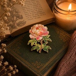 May include: A gold-toned metal brooch shaped like a blooming peony flower with layered pink and white petals and green leaves. The design features intricate gold outlines separating the coloured enamel sections, creating a detailed floral botanical accessory.