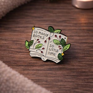 May include: Enamel pin depicting an open book with the text "SO MANY BOOKS" and "SO LITTLE TIME." The pin features green leaves and red berries. The pin is set on a wooden surface.