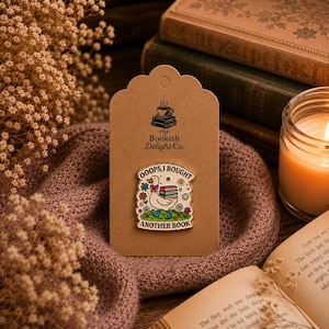 May include: A white enamel pin featuring a cartoon duck wearing blue boots and carrying a stack of books, surrounded by colourful flowers and stars. The text reads OOOPS, I BOUGHT ANOTHER BOOK. The pin is attached to a brown card with the logo THE BOOKISH DELIGHT CO. and a small stack of books with a steaming cup on top.