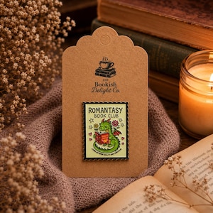 Peut inclure: Une broche émaillée rectangulaire représentant un dragon vert lisant un livre, avec le texte "ROMANTASY BOOK CLUB" et "LIVING BETWEEN REALITY AND FANTASY" sur fond jaune pâle, fixée sur une carte brune marquée "The Bookish Delight Co."