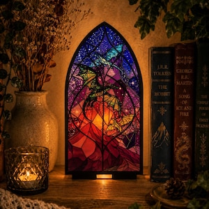 May include: A pointed arch-shaped lamp featuring a stained glass design of two dragons flying against a starry night sky and a glowing sunset over mountains. The lamp has a black frame and emits a warm light from behind the glass panel.