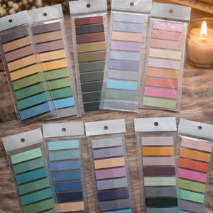 May include: A collection of colorful sticky note pads in various shades, including greens, blues, oranges, and purples. Each pad is packaged in a clear plastic sleeve. The pads are arranged on a textured surface.