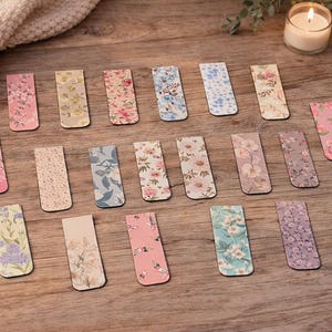 May include: A selection of rectangular bookmarks with floral designs in shades of pink, yellow, blue, and purple. The bookmarks are displayed on a wooden surface, with a lit candle and foliage in the background.