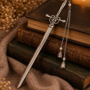 May include: A silver-toned metal hair accessory shaped like a miniature sword with a detailed Celtic knot crossguard. The hilt features a dangling silver chain adorned with two small white pearls and two metallic bell-shaped charms at the ends.