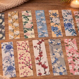 May include: A selection of floral bookmarks. Each bookmark displays a unique floral pattern, such as white magnolias, pink cherry blossoms, and blue peonies. The rectangular bookmarks are arranged on a wooden surface.