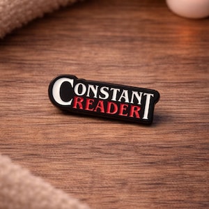May include: A black and red enamel pin with the text "CONSTANT READER" in white lettering. The pin is oval-shaped and rests on a wooden surface. A lit candle and a soft, beige fabric are in the background.