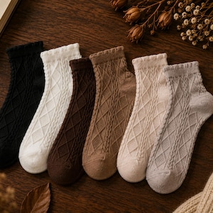 May include: A set of six ankle socks in black, white, dark brown, tan, cream, and light grey. Each sock features a textured diamond cable knit pattern and a ribbed cuff. The socks are arranged in a row, showcasing the variety of neutral colours.