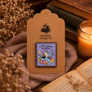 May include: A rectangular enamel pin shaped like a postage stamp with a light purple border. The design features a white duck wearing a floral blanket, reading a pink book with a flashlight under a yellow crescent moon and stars, labelled Late Night Reader Club.