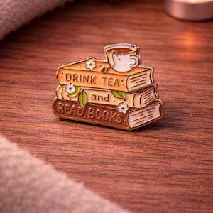 May include: A gold enamel pin depicting a stack of books with the text "DRINK TEA" and "READ BOOKS". A teacup sits on top. The pin is decorated with white flowers and green leaves. The pin is on a wooden surface.