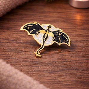 May include: A gold-colored dragon pin with black wings and a white background. The dragon has a long tail and is set against a white circle. The pin is on a wooden surface.