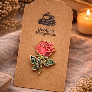 May include: A rose-shaped enamel pin with red petals, green leaves, and gold detailing, presented on a brown tag featuring "The Bookish Delight Co." logo. The pin is set against a soft, neutral-toned backdrop.