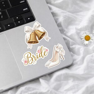 May include: A set of three stickers featuring two gold wedding bells tied with a white ribbon, the word Bride in gold cursive script decorated with small pink and white flowers, and a pair of white high-heeled shoes with a white bow.
