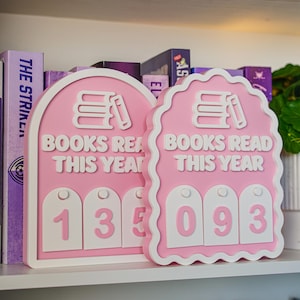 May include: Two pink and white arched signs with the text BOOKS READ THIS YEAR. The left sign has a stack of books icon and the number 135 on white tags. The right sign has a scalloped edge, a stack of books icon, and the number 093 on white tags.