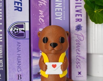 May include: A brown otter figurine with a textured surface, wearing a bright yellow vest and holding a white envelope featuring a small red heart icon on the front. The otter has large black eyes and a black nose, standing upright.