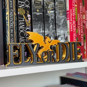 May include: A 3D-printed decorative sign featuring the text FLY OR DIE in black lettering with gold edges, accented by a gold dragon silhouette positioned above the word OR, standing on a white shelf in front of several books.