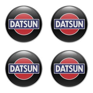 Può includere: Quattro adesivi lucidi neri con l'emblema Datsun. Ogni emblema presenta un cerchio rosso, un rettangolo bianco con la scritta "DATSUN" in blu e un bordo nero. Gli adesivi sono rotondi e hanno una superficie riflettente.