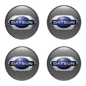 May include: Four Datsun logo emblems. Each emblem features the Datsun logo in a blue and silver oval shape, set against a carbon fiber background. The emblems are circular and have a glossy finish.