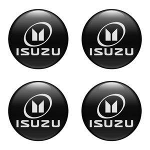 May include: Four black, glossy Isuzu emblem stickers. Each circular sticker features a white stylized 'M' within a white oval, with the word 'ISUZU' in white block letters below. The stickers are arranged in a square formation.