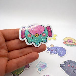 Op de afbeelding: Een holografische sticker met een cartoon olifant met een turquoise lichaam, roze oren en een roze en glinsterende kop. De olifant heeft een kleine gele cirkel in het midden. Andere stickers van zeedieren staan op de achtergrond.