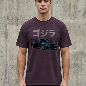 May include: Dark purple t-shirt featuring a blue sports car graphic with the Japanese word "ゴジラ" above it. The shirt is a crew neck and has a vintage look.