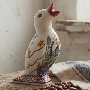 May include: A ceramic bird figurine with a cream-colored base, featuring a bright red beak and a decorative pattern of blue and green leaves with small orange flowers. The base of the bird is adorned with a geometric red and blue pattern.