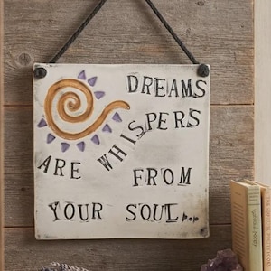 May include: A square white wall sign with black text that reads DREAMS ARE WHISPERS FROM YOUR SOUL. The sign features a brown spiral graphic with purple accents and is suspended by a black rope from a metal hook on a wooden wall.