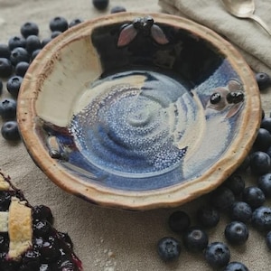 May include: A round ceramic bowl with a glazed finish in shades of deep blue, cream, and brown. The bowl features a textured rim and a smooth, glossy interior. It is surrounded by scattered fresh blueberries and a slice of blueberry pie.