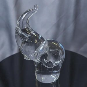 May include: A clear glass figurine of an elephant standing with its trunk raised upward. The sculpture is crafted from transparent, smooth glass, capturing the distinct shape of the elephant's head, ears, and body in a reflective, solid form.