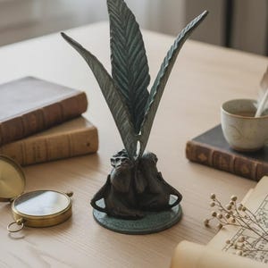 May include: A bronze-colored decorative sculpture of stylized feathers on a round base. The sculpture is on a wooden table, with books, a compass, and a teacup in the background. The sculpture is approximately 8 inches tall.