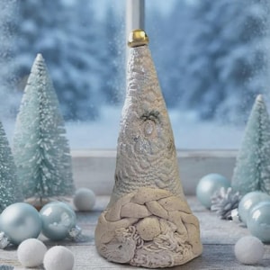 May include: A cone-shaped gnome figurine with a textured white hat topped with a small gold bead, a braided beard, and a round nose. The gnome is surrounded by light blue ornaments, white pom-poms, silver pinecones, and miniature bottle brush trees.