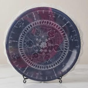 May include: A circular decorative plate featuring a deep purple and blue gradient background with intricate white line art. The design includes concentric geometric patterns, stylized floral motifs, and a central star-like element, all displayed on a black metal stand.