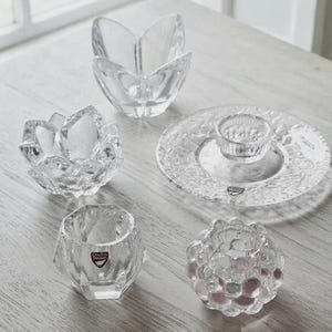 May include: A collection of five clear glass items including two flower-shaped bowls, one small faceted bowl with a label, one textured round candle holder, and one circular plate with a central raised holder, all featuring Orrefors Sweden branding.