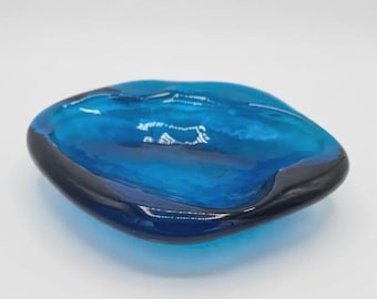 May include: A small, shallow bowl crafted from translucent blue glass. The dish features an irregular, organic shape with a smooth, polished surface and a deep cobalt blue rim that transitions into a lighter, vibrant turquoise center.