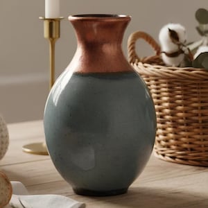 May include: A two-toned ceramic vase with a rounded body and a flared neck. The upper portion is a copper color, while the lower part is a muted blue-gray. The vase is displayed on a wooden surface, with a candle holder and a woven basket in the background.