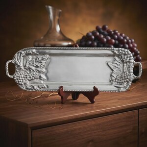 May include: A rectangular silver-toned metal serving tray featuring embossed grape and vine leaf motifs on both ends. The tray has integrated handles on each side and a rope-style border detail, presented on a dark brown display stand.