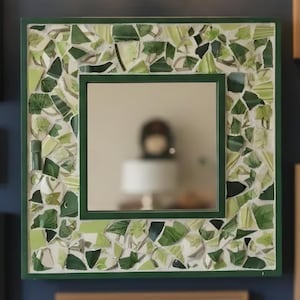 May include: A square wall mirror featuring a frame decorated with a mosaic of various green and white glass shards. The central reflective glass is surrounded by a dark green border, with the mosaic pieces set in a light-colored grout.