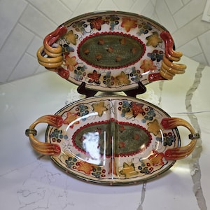 May include: Two oval ceramic serving platters with handles. The platters have a cream-colored background with a green and red oval center. The edges are decorated with colorful leaves and grapes. The handles are a twisted design.