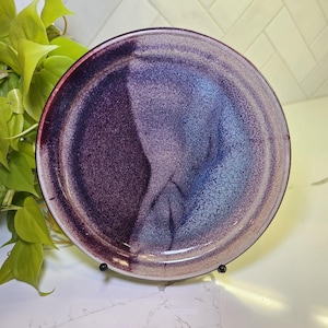 May include: A round ceramic plate with a speckled, glazed finish. The plate features a gradient of colors, transitioning from deep purple to light blue. The rim is a darker shade of purple.