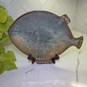 May include: A ceramic serving dish in the shape of a fish. The dish has a blue and tan glaze with a dark brown rim. It is displayed on a brown stand. The fish-shaped dish has fins and a tail.