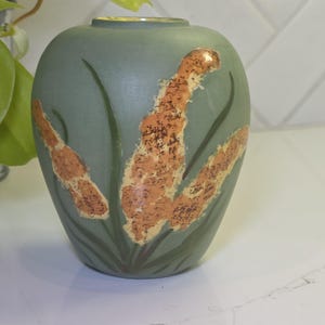 May include: A teal-colored ceramic vase with a rounded shape. The vase is decorated with orange and yellow cattails and green leaves. The vase has a smooth, matte finish and a small opening at the top.