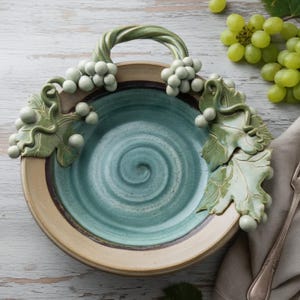 May include: A ceramic serving dish with a teal interior and a beige rim. The dish features a handle and grape vine accents in green. The handle is a twisted vine shape, and the rim has a spiral design. The dish is designed for serving.