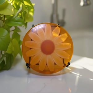 May include: A round, amber-colored decorative piece featuring a large, detailed daisy with yellow petals and an orange center. The piece is displayed on a black metal stand, showcasing the floral design. The background is a white surface.