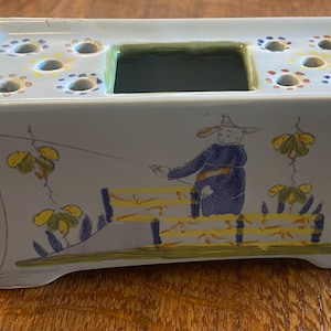 May include: A rectangular ceramic flower vase with a light blue exterior. The vase features a painted scene of a person fishing, with yellow and blue accents. The top has several holes for arranging flowers, and a central square opening.