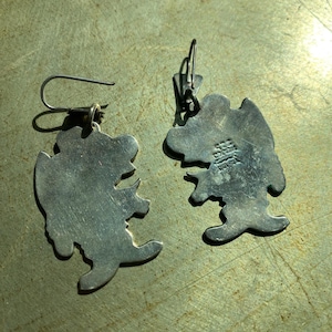 Op de afbeelding: Een paar zilverkleurige oorbellen. Elke oorbel heeft een gestileerde beerachtige figuur met een haak en sluiting. De oorbellen worden gepresenteerd op een getextureerd, metalen oppervlak.