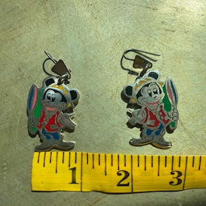 Op de afbeelding: Een paar zilverkleurige Mickey Mouse-oorbellen. Elke oorbel heeft een kleurrijk Mickey Mouse-ontwerp met een surfboard. De oorbellen hebben een haaksluiting en zijn geplaatst tegen een getextureerde achtergrond.