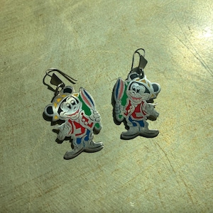 Op de afbeelding: Een paar zilverkleurige oorbellen met een cartoonkarakterontwerp. De oorbellen tonen een personage in een rode jas, blauwe broek en een gele hoed, die een kleurrijk object vasthoudt. De oorbellen hebben een haaksluiting.