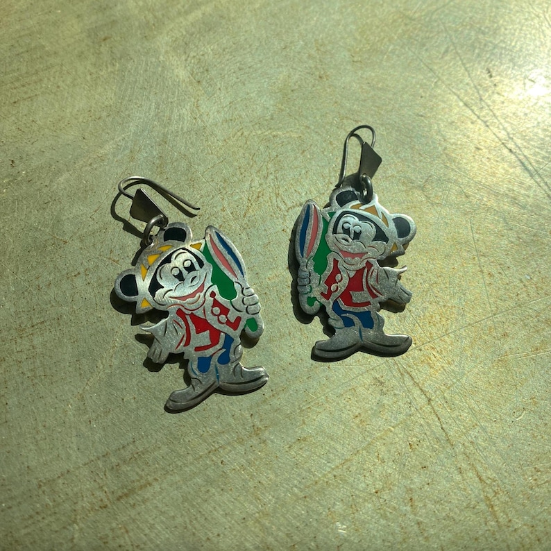 Op de afbeelding: Een paar zilverkleurige oorbellen met een cartoonkarakterontwerp. De oorbellen tonen een personage in een rode jas, blauwe broek en een gele hoed, die een kleurrijk object vasthoudt. De oorbellen hebben een haaksluiting.
