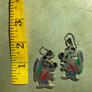 Op de afbeelding: Een paar zilverkleurige Mickey Mouse oorbellen. Elke oorbel heeft een kleurrijk emaille ontwerp van Mickey Mouse met een surfplank. De oorbellen hebben een haaksluiting en zijn ongeveer 2,5 cm hoog.