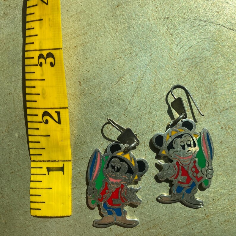 Op de afbeelding: Een paar zilverkleurige Mickey Mouse oorbellen. Elke oorbel heeft een kleurrijk emaille ontwerp van Mickey Mouse met een surfplank. De oorbellen hebben een haaksluiting en zijn ongeveer 2,5 cm hoog.