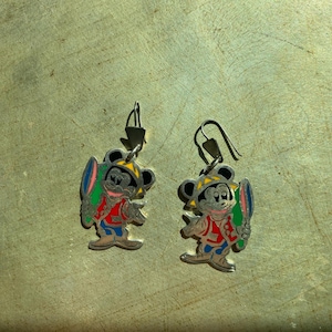 Op de afbeelding: Een paar zilverkleurige oorbellen met een cartoon muis karakter. De muis draagt een gele hoed, een rode jas, een blauwe broek en houdt een kleurrijk surfboard vast. De oorbellen hebben een haaksluiting.